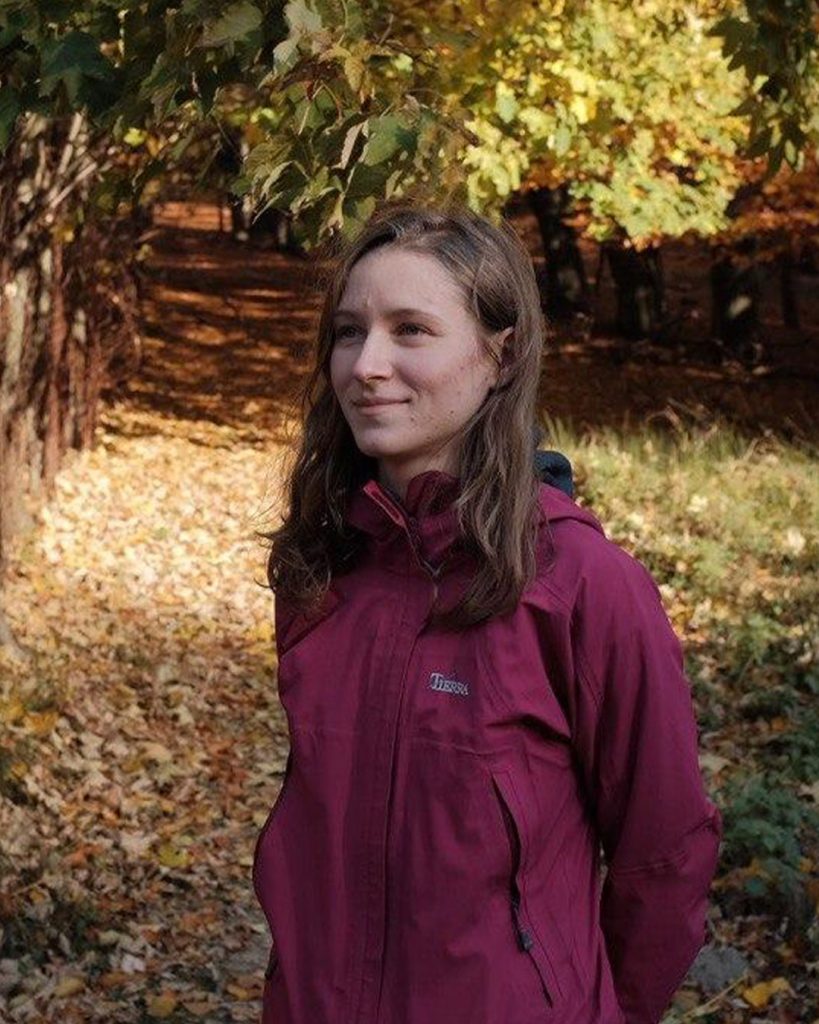Coach Alina in a maroon jacket standing on a leafy path in an autumn forest, looking thoughtful.