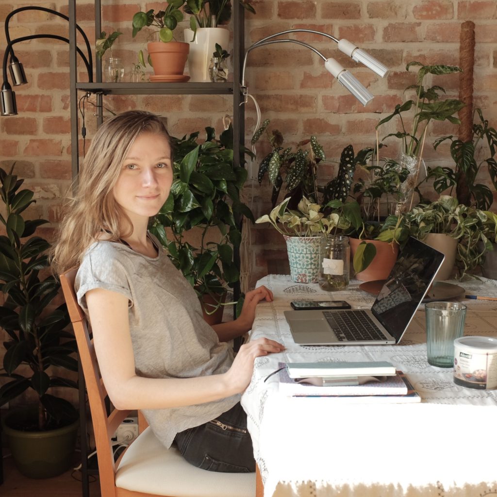 Coach Alina sitting at a table with a laptop, surrounded by lush green plants, working in a cozy home office with a brick wall in the background.